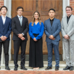 Photo of five students in dress clothes in front of a red brick building and wooden doors. A woman stands in the center flanked by two men on both sides of her.